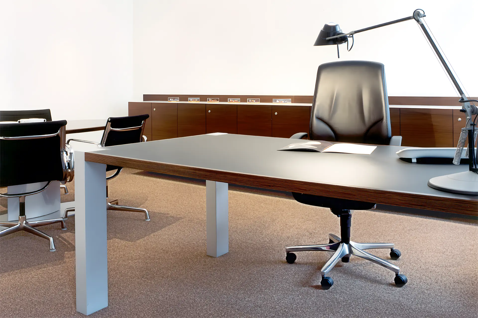 Hinter einen Arbeitstisch mit Tischleuchte und Bürostuhl befindet sich ein Sideboard in dunkel gebeiztem amerikanischem Nussbaum.