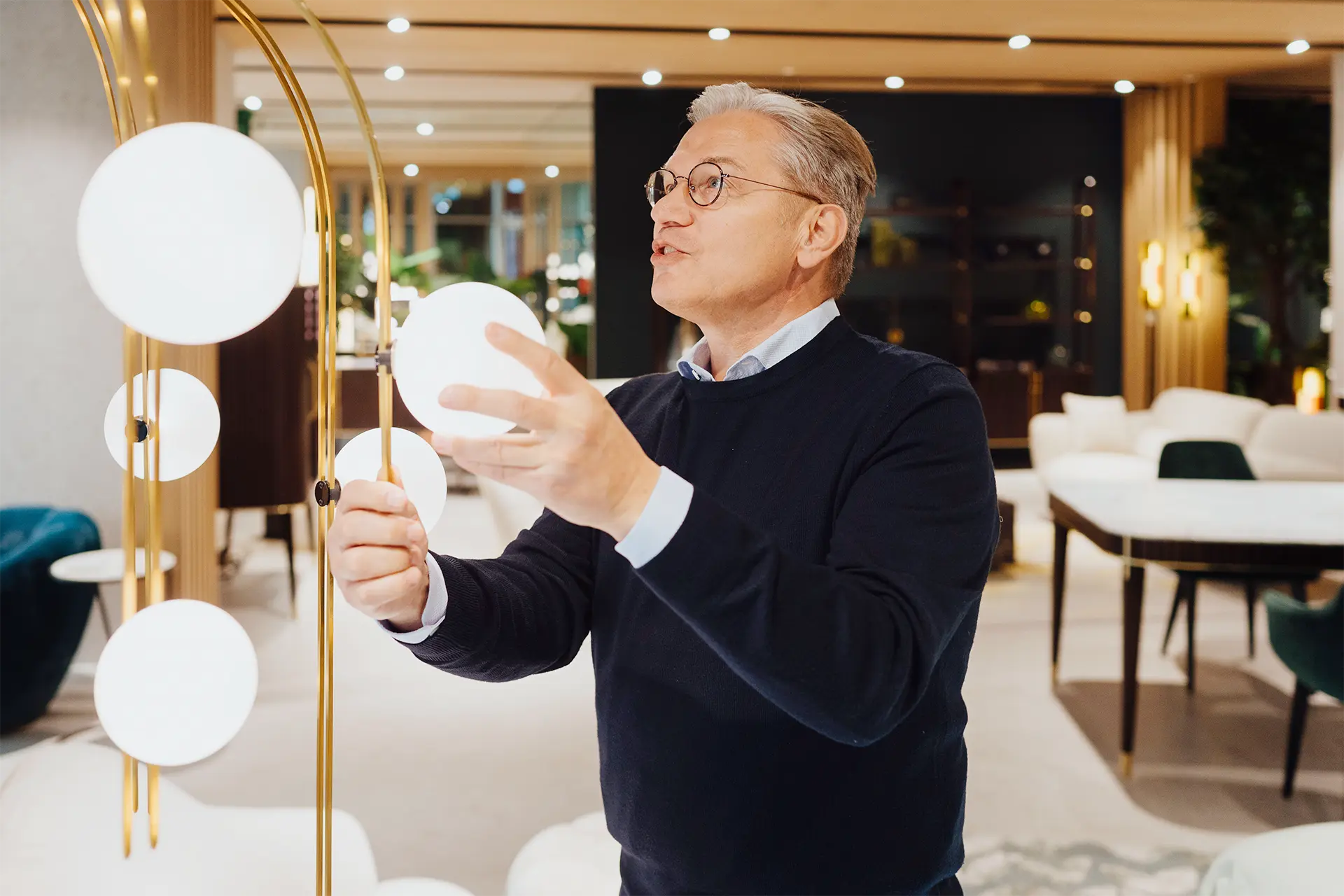 Ulrich Weinkath – Besuch in einem Showroom am Neuen Wall – Begutachtung Stehlampe Ulrich Weinkath steht in einem Interior-Showroom am Neuen Wall. Er begutachtet eine Stehlampe mit kreisrunden Lampenschirmen aus Glas.
